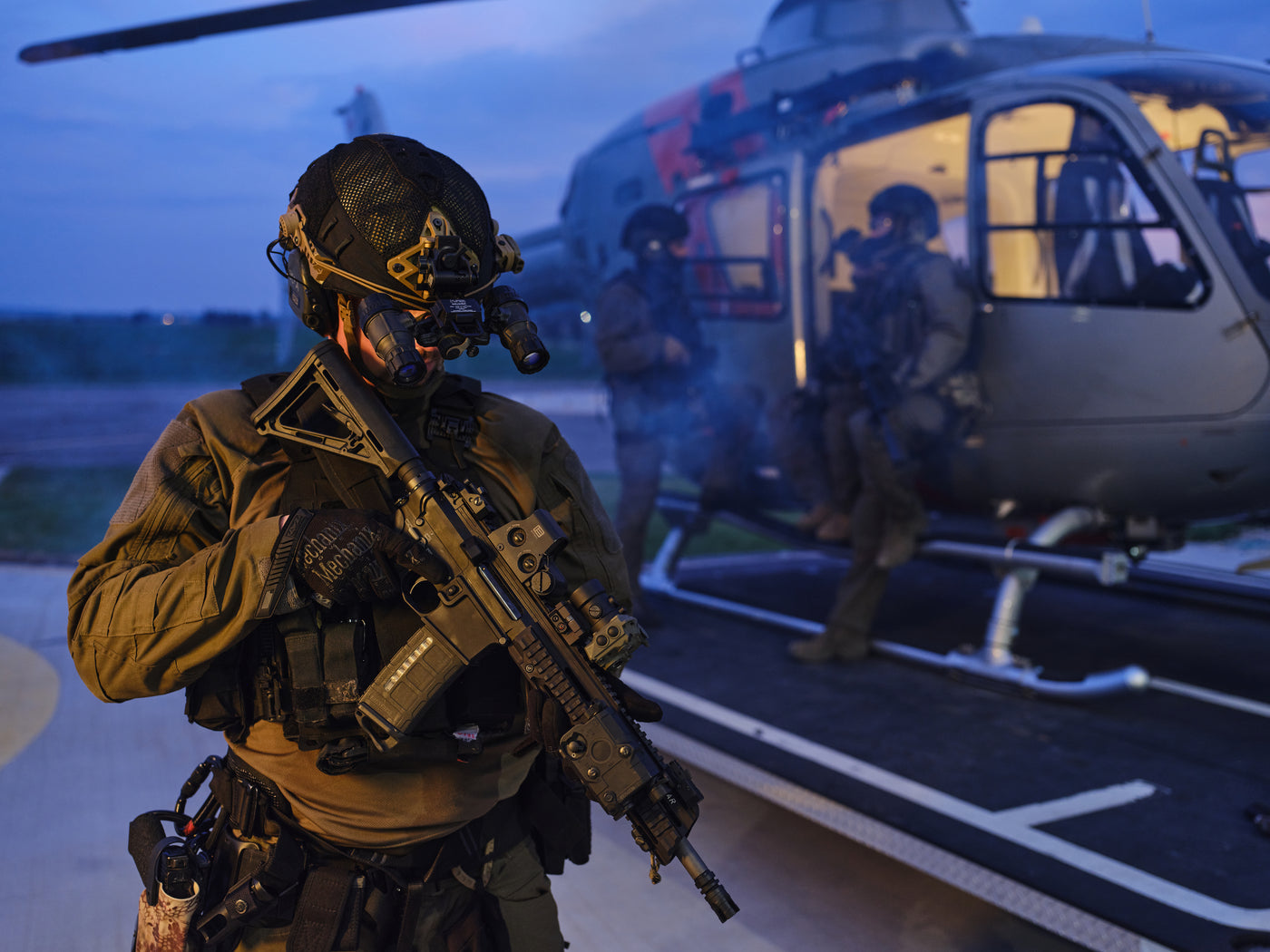 Mehrere Streitkräfte stehen und sitzen vor einem Helikopte. Im Vordergrund hat die Person eine TILO auf ihrer Waffe montiert.